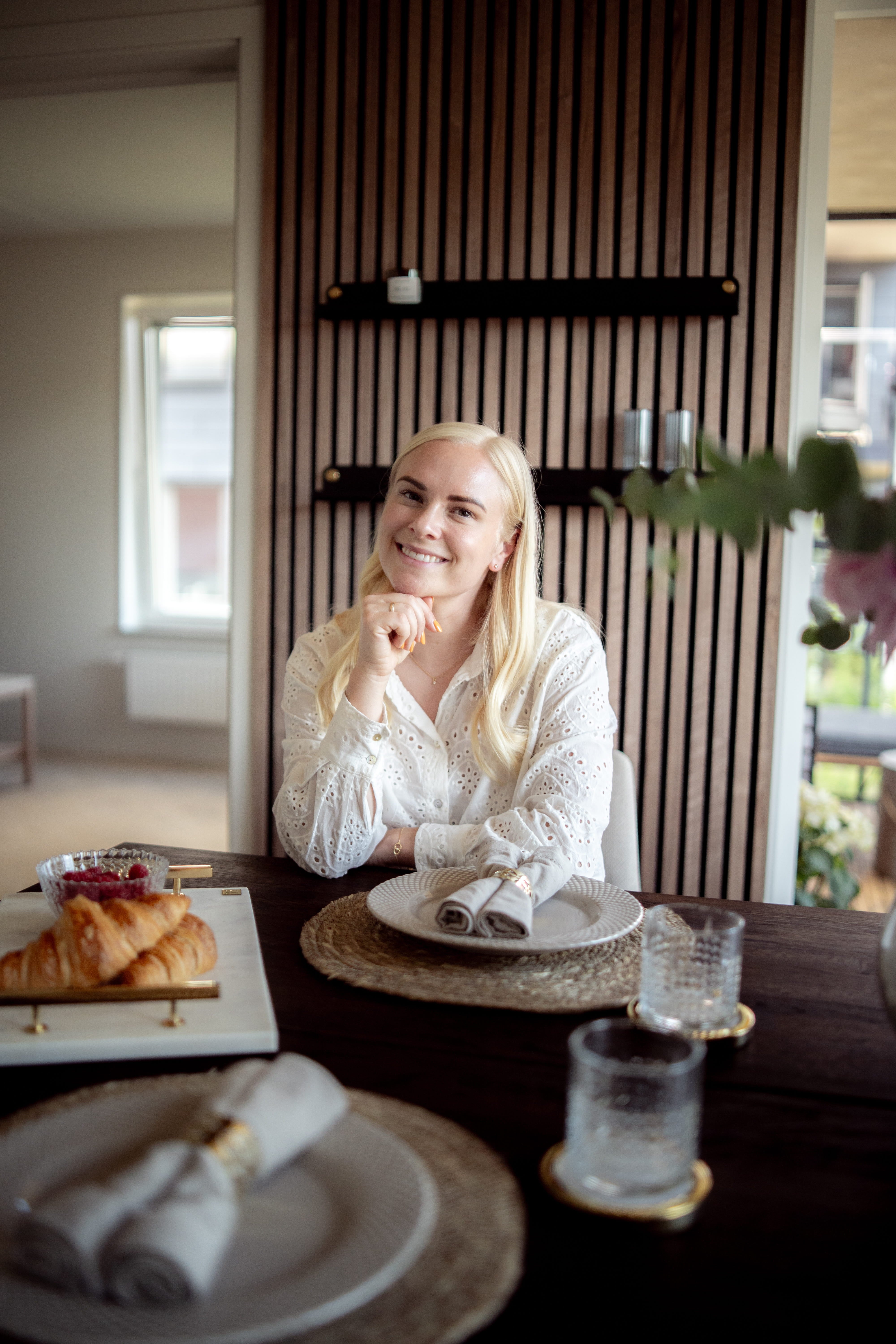 Een vrouw met lang blond haar zit aan een ontbijttafel met croissants en glaswerk, naast een Scandinavisch akoestisch paneel aan de muur.